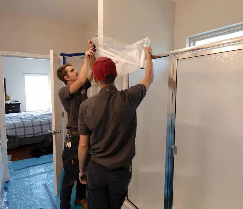 Two workers in uniforms cover a bathroom doorway with plastic sheeting, preparing the area next to a shower for maintenance or renovation.