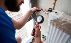 A person in a blue shirt checks a radiator's pressure with a gauge. The white radiator is partly wrapped in plastic, indicating recent installation or repair.