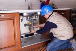 A plumber in a blue hard hat and beige vest kneels on a striped rug, fixing pipes under a kitchen sink in a tiled and wooden cabinet setting.