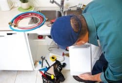 A plumber in a blue shirt and cap examines under a kitchen sink with tools and hoses nearby. He holds a clipboard while evaluating the repair.
