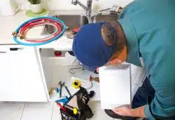 A plumber in a blue shirt and cap examines under a kitchen sink with tools and hoses nearby. He holds a clipboard while evaluating the repair.