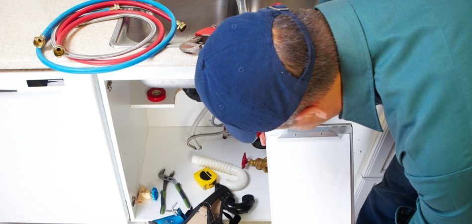 A plumber in a blue shirt and cap examines under a kitchen sink with tools and hoses nearby. He holds a clipboard while evaluating the repair.