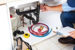 A plumber inspects under-sink pipes, surrounded by tools like a tape measure, wrench, and clipboard. Nearby are blue and red hoses, conveying maintenance work.