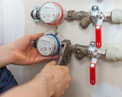 Close-up of hands using a wrench to adjust water meters with red and blue rims, attached to pipes with red-handled valves, indicating plumbing maintenance.