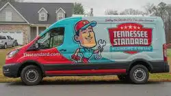 A Tennessee Standard Plumbing & Drain service van is parked outside a house, featuring company branding and a cartoon plumber ready to fix issues like improperly slope drain pipes with his wrench and toolbox.