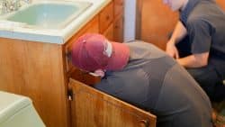 Two people in work uniforms inspect plumbing under a bathroom sink with wooden cabinets.