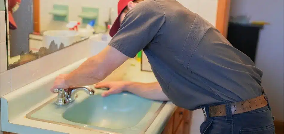 A person wearing a red cap and work uniform is fixing or inspecting a bathroom sink faucet, possibly addressing sudden drops in water pressure.