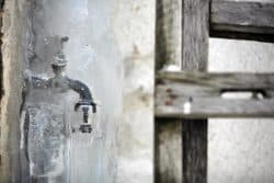 A water faucet encased in a thick layer of ice next to a weathered wooden structure highlights common plumbing risks during power outages.