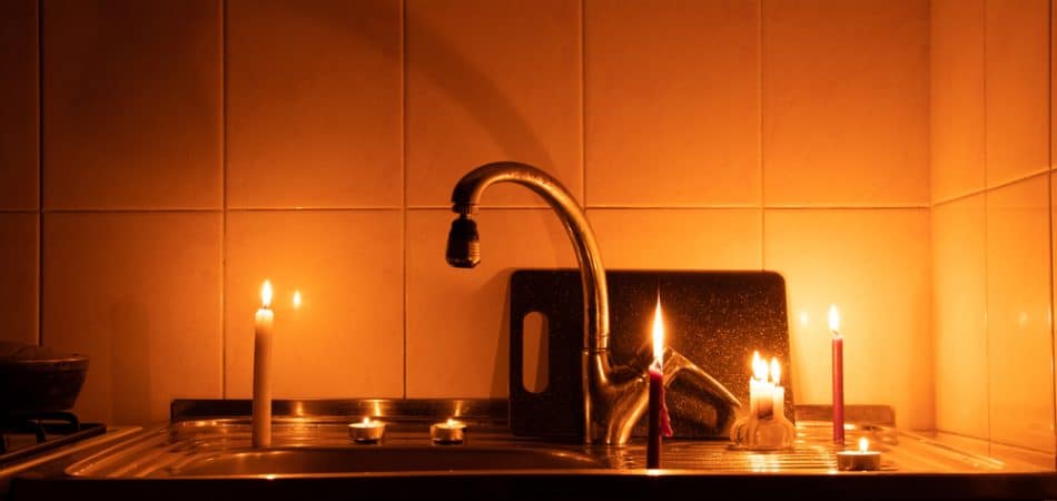 A kitchen sink lit by two candles with a paper displaying a sad face propped against the faucet; the dimly lit setting hints at plumbing risks during power outages.