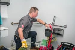 A plumber in uniform kneels in a bathroom, operating a drain cleaning machine with tools and equipment nearby.