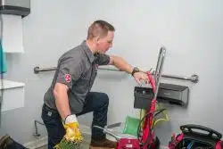 A plumber in uniform kneels in a bathroom, operating a drain cleaning machine with tools and equipment nearby.