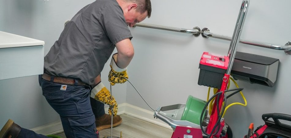 A plumber kneels on the floor using a drain cleaning machine to tackle plumbing problems and clear a pipe in a bathroom. Tools and equipment are nearby.