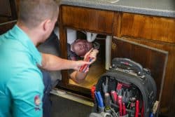 A plumber lies under a kitchen sink, using a wrench on the pipes, while another person hands him a tool. A tool bag sits nearby.