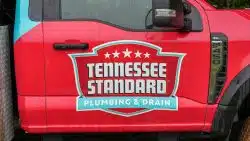 Red truck door with a "Tennessee Standard Plumbing & Drain" logo, featuring a shield design and stars, is shown in the rain, highlighting their trusted plumbing inspection services.