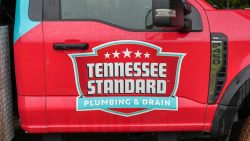 Red truck door featuring the "Tennessee Standard Plumbing & Drain" logo, a shield with four white stars, and specialized in commercial drain cleaning.