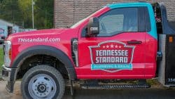 Red utility truck with "Tennessee Standard Plumbing & Drain" logo and website "TNStandard.com" on the door, parked on a street beside a brick building, ready to handle plumbing issues like high water pressure.