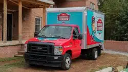 A red and blue Tennessee Standard Plumbing & Drain box truck is parked outside a brick house near a porch and some trees, ready to help homeowners tackle plumbing issues after renovations.