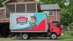 A red and teal Tennessee Standard Plumbing & Drain truck is parked in a driveway beside a brick house with a raised wooden porch, ready to service drainage systems in Knoxville.