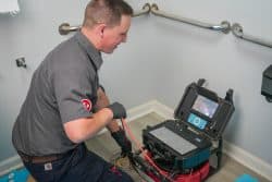 A plumber uses advanced plumbing tools, including a drain auger machine, to clear a clogged pipe in a restroom, kneeling next to an exposed drain with equipment nearby for improved repair accuracy.