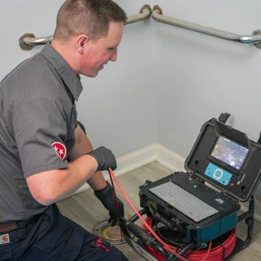 A plumber uses advanced plumbing tools, including a drain auger machine, to clear a clogged pipe in a restroom, kneeling next to an exposed drain with equipment nearby for improved repair accuracy.