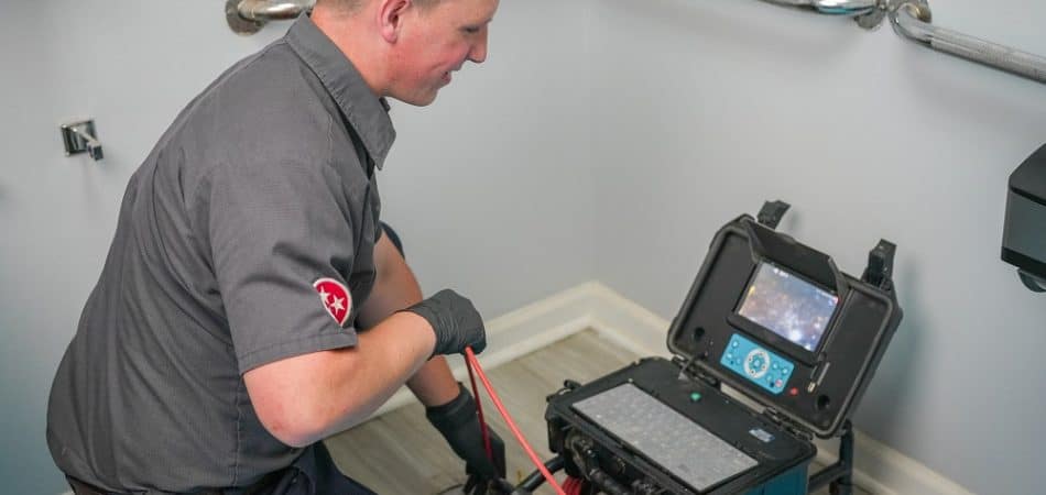 A plumber uses advanced plumbing tools, including a drain auger machine, to clear a clogged pipe in a restroom, kneeling next to an exposed drain with equipment nearby for improved repair accuracy.