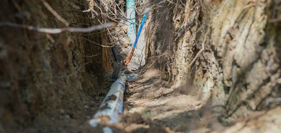 A close-up view of underground utility pipes and cables running through a narrow trench in the soil.