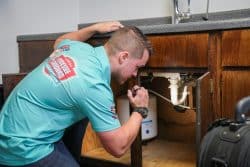 A person wearing a blue shirt uses a flashlight to inspect plumbing under a kitchen sink cabinet, checking for plumbing issues after renovations.
