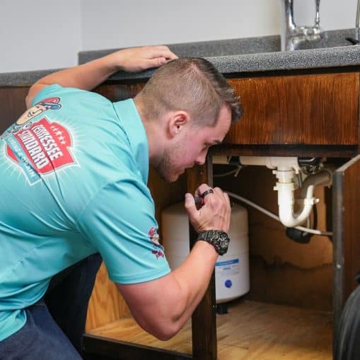 A man in a blue shirt uses a flashlight to inspect plumbing under a kitchen sink.