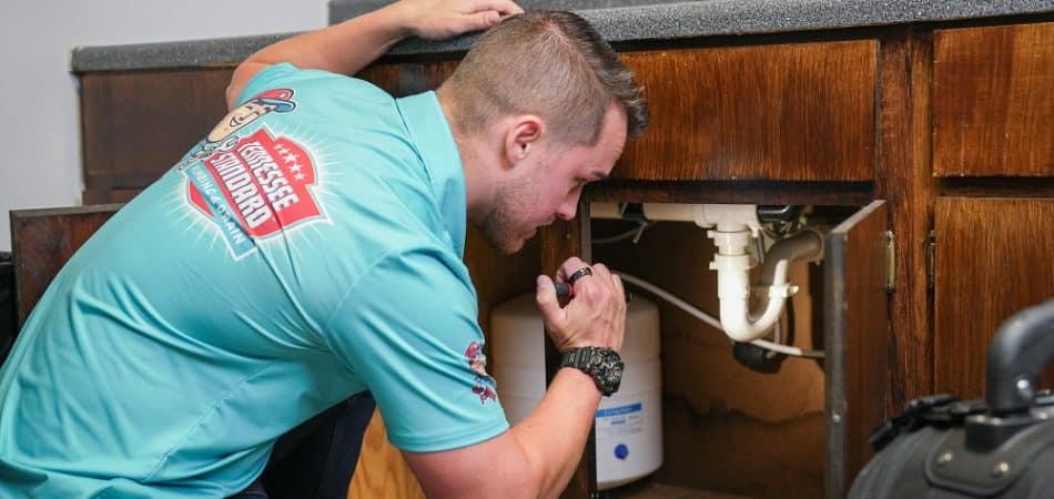 A man in a blue shirt uses a flashlight to inspect plumbing under a kitchen sink.