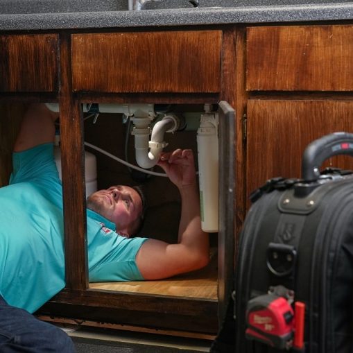 A man in a blue shirt lies under a kitchen sink, fixing plumbing