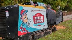 A worker operates a tracked machine with "Tennessee Standard Plumbing & Drain" branding on the side, parked on grass near a red pickup truck.