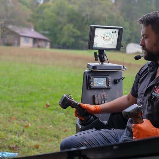 A man wearing orange gloves and a headset operates heavy machinery using dual control sticks, with a monitor displaying data in front of him—demonstrating plumbing innovation in an outdoor rural setting.