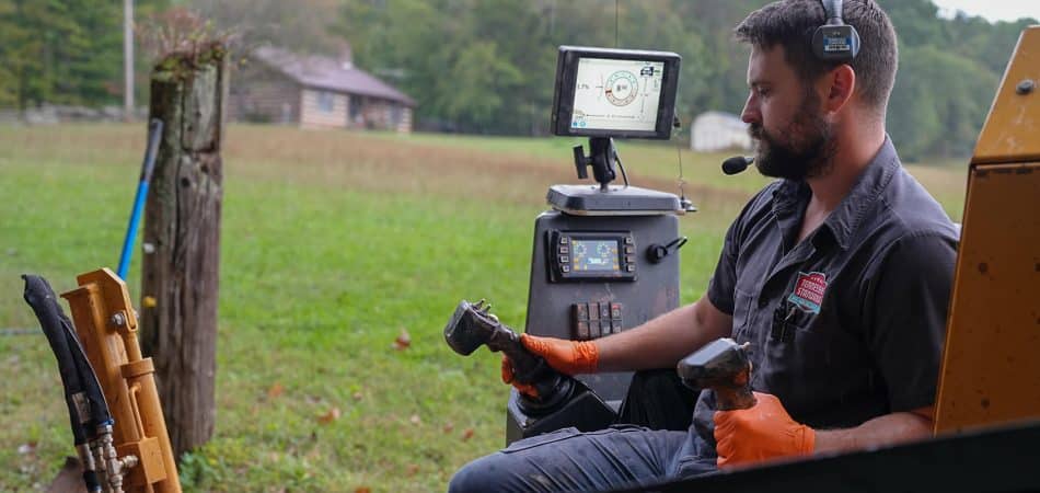 A man wearing orange gloves and a headset operates heavy machinery using dual control sticks, with a monitor displaying data in front of him—demonstrating plumbing innovation in an outdoor rural setting.