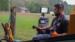 A man wearing gloves and a headset operates machinery using joysticks, with control screens and a grassy field in the background, showcasing how advanced plumbing tools improve repair accuracy.