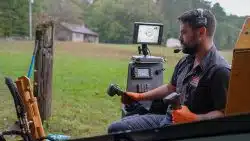 A man wearing gloves and a headset operates machinery using joysticks, with control screens and a grassy field in the background, showcasing how advanced plumbing tools improve repair accuracy.