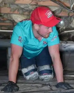 A person wearing knee pads, gloves, a red cap, and a turquoise shirt is crawling on hands and knees inside an unfinished attic or crawl space with exposed insulation, possibly inspecting for Backflow Testing in Knoxville.