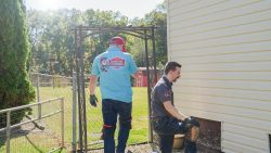 Two men wearing work clothes and knee pads work outside near a house—one kneels by the foundation, possibly assessing Sewer Line Repair vs. Replacement, while the other walks through a gate.