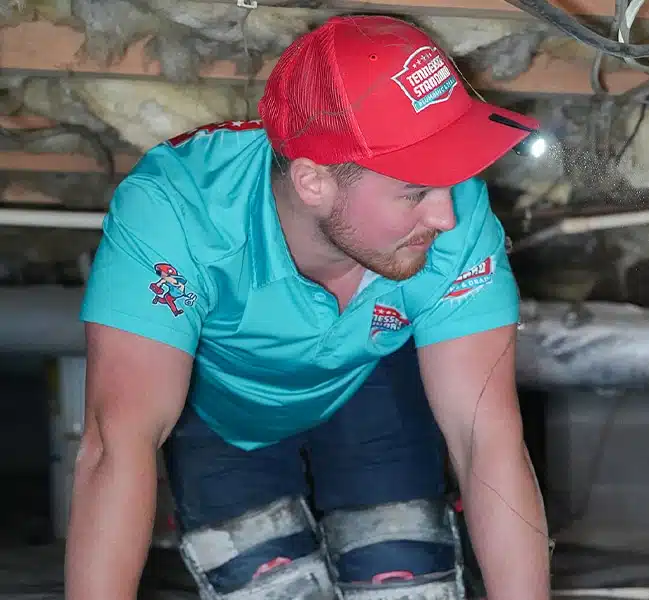 A plumber crawling in a space with exposed insulation and wooden beams above him.