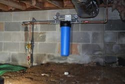 A blue whole-house water filter system is mounted on a basement wall, connected to copper pipes with gauges and valves.