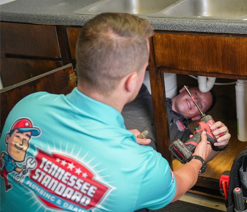 Two plumbers work under a kitchen sink; one holds a power drill while the other lies inside the cabinet. The visible shirt reads