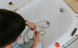 A man wearing work overalls uses a plunger to unclog a bathtub drain in a tiled bathroom.
