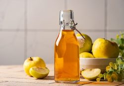 A glass bottle of apple cider vinegar, often used for natural drain cleaning in Knoxville, sits on a counter next to a white bowl filled with whole and sliced apples.