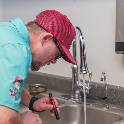 A person wearing a teal shirt and red cap inspects a sink with running water using a flashlight in a kitchen or utility room.