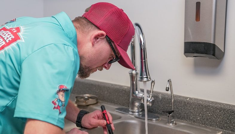 A person wearing a teal shirt and red cap inspects a sink with running water using a flashlight in a kitchen or utility room.