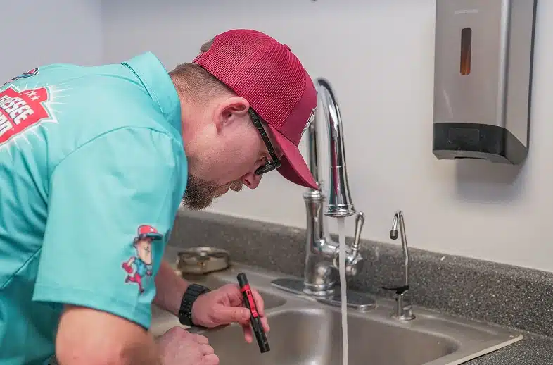 A person wearing a teal shirt and red cap inspects a sink with running water using a flashlight in a kitchen or utility room.