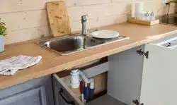 A kitchen counter with a stainless steel sink, faucet, cutting board, plate, dish towel, and an open cabinet showing exposed plumbing underneath.