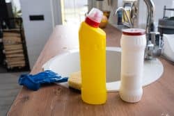 Two cleaning product bottles, a sponge, and a blue glove are placed on a kitchen counter next to a sink.