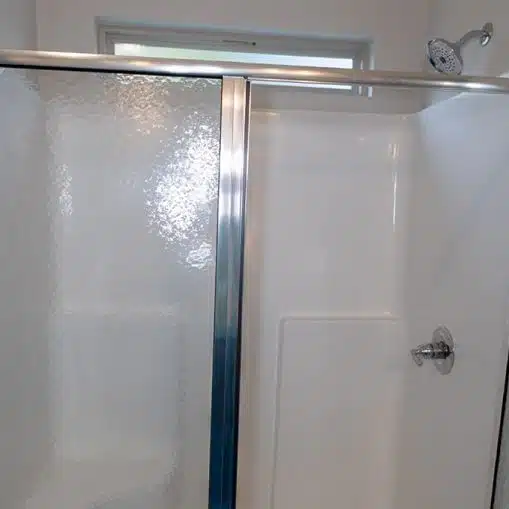 Glass shower with sliding door, chrome frame, and white fiberglass walls; showerhead and control handle visible on the right.