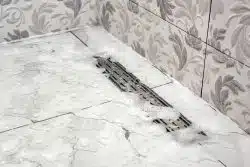 Soapy water on a tiled bathroom floor drains through a rectangular metal grate near the corner, with decorative leaf-patterned wall tiles visible.