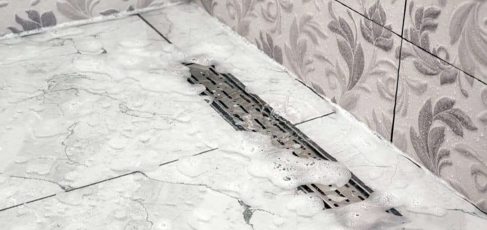 Soapy water on a tiled bathroom floor drains through a rectangular metal grate near the corner, with decorative leaf-patterned wall tiles visible.